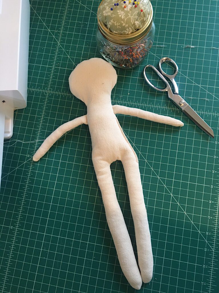 Cloth Doll Body laying on sewing table with fabric scissor and sewing pins